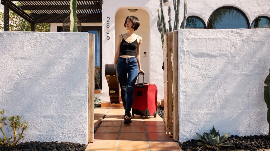 Image of a woman walking out of a home carrying an acoustic guitar in a case and wheeling a suitcase