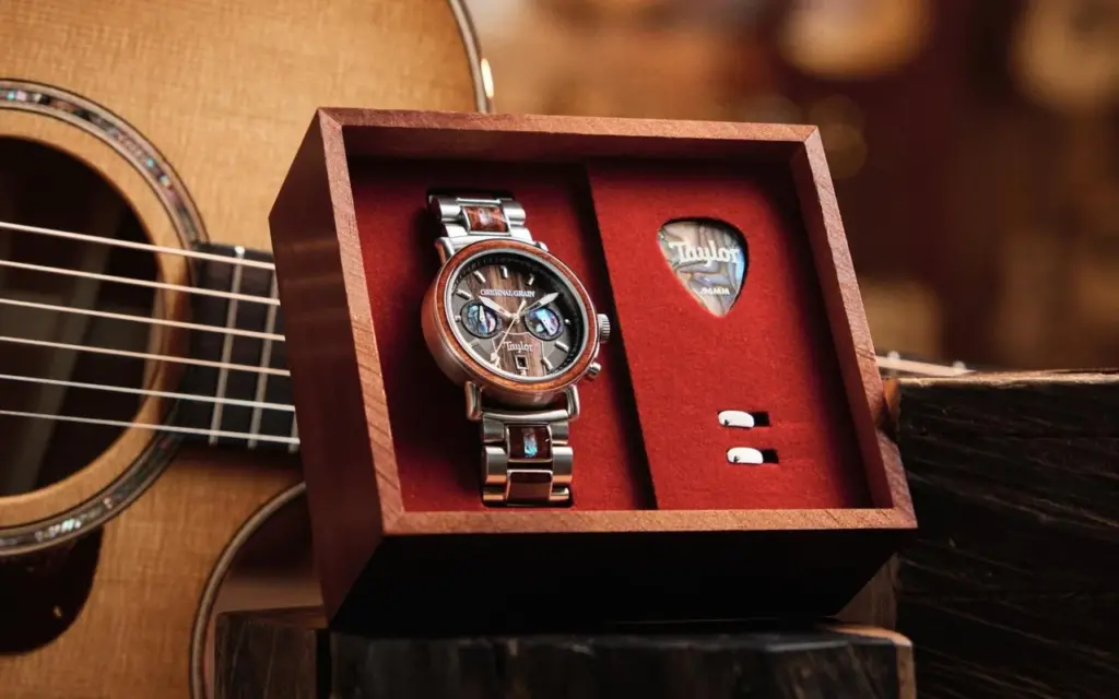 a wristwatch made by Original Grain in a red-lined wooden box with a guitar pick sits in front of a Taylor acoustic guitar