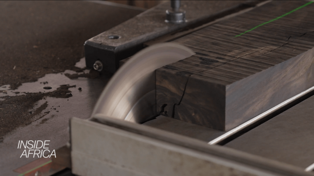 A table saw cuts through a large block of ebony wood in a mill with the logo for CNN's Inside Africa in the foreground